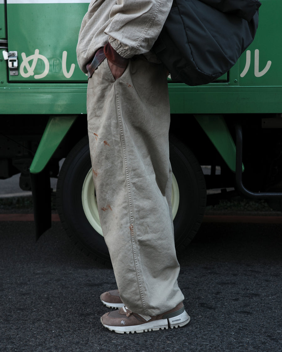 フロントに2タックを施し、ワタリから裾にかけてゆとりを持たせたワイルドなワイドシルエット。裾のドローコードでシルエットの変化も自在に楽しめます。素材には耐久性に優れ、綾織特有の柔らかさを持つ厚手チノクロスを採用。ストーンバイオウォッシュと汚し加工を重ねることで、長年穿き込んだようなリアルな質感と深みのある表情を引き出しました。

Wild, wide-silhouette pants with double-tucked fronts and adjustable hem drawcords. Crafted from durable, soft chino cloth, this BEIGE pair features a stone bio-wash and staining process. This creates an authentic, rugged texture and a deeply weathered look, blending vintage workwear aesthetics with a modern, voluminous fit.